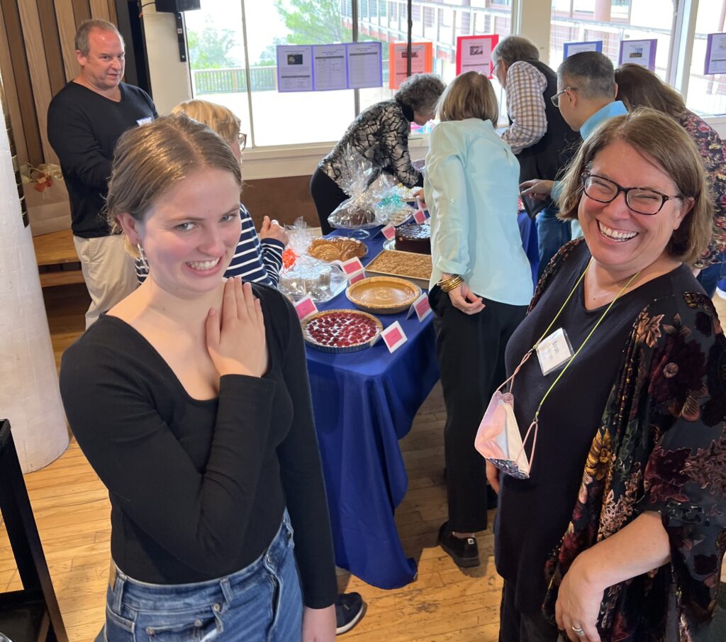 People gathered around a table full of desserts with two happy ladies in the forefront smiling at the camera.